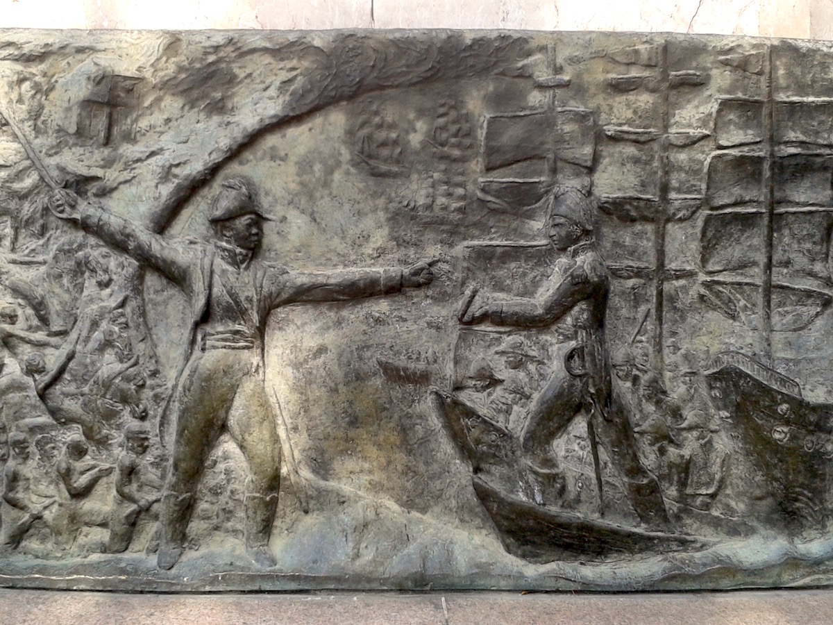 Sculpture of a man on a ship being greeted by another man with a sword.