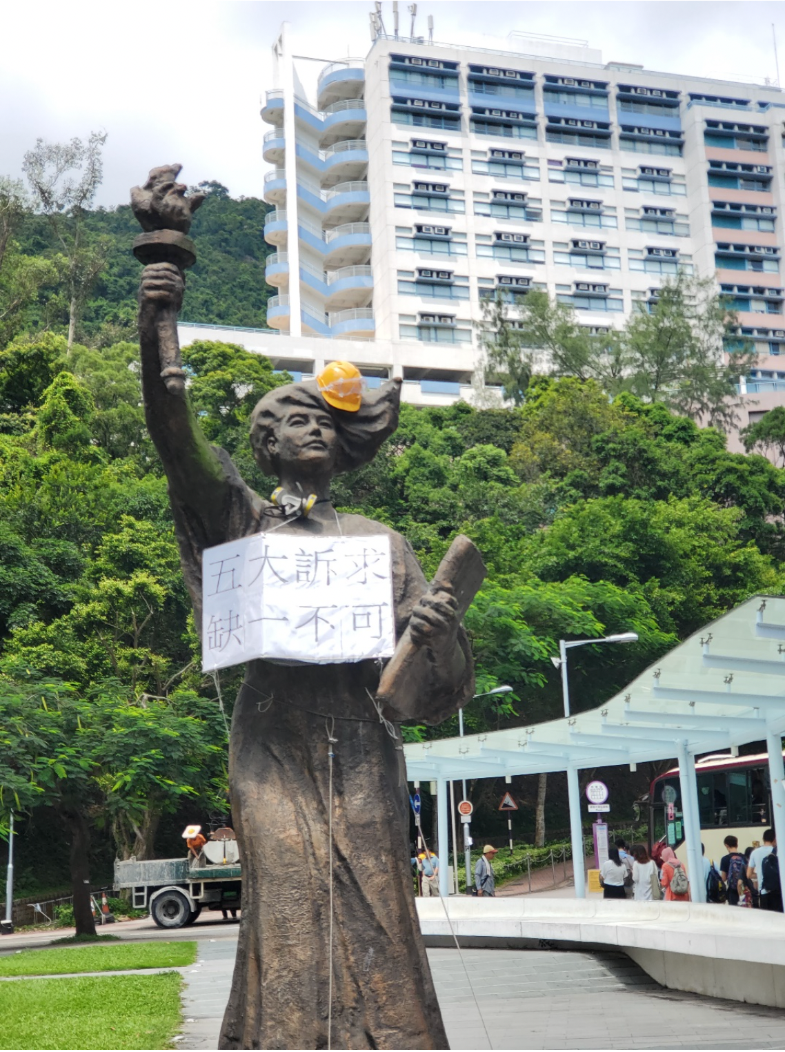 Statue wearing a worker helmet and a cardboard sign around her neck with Chinese characters.